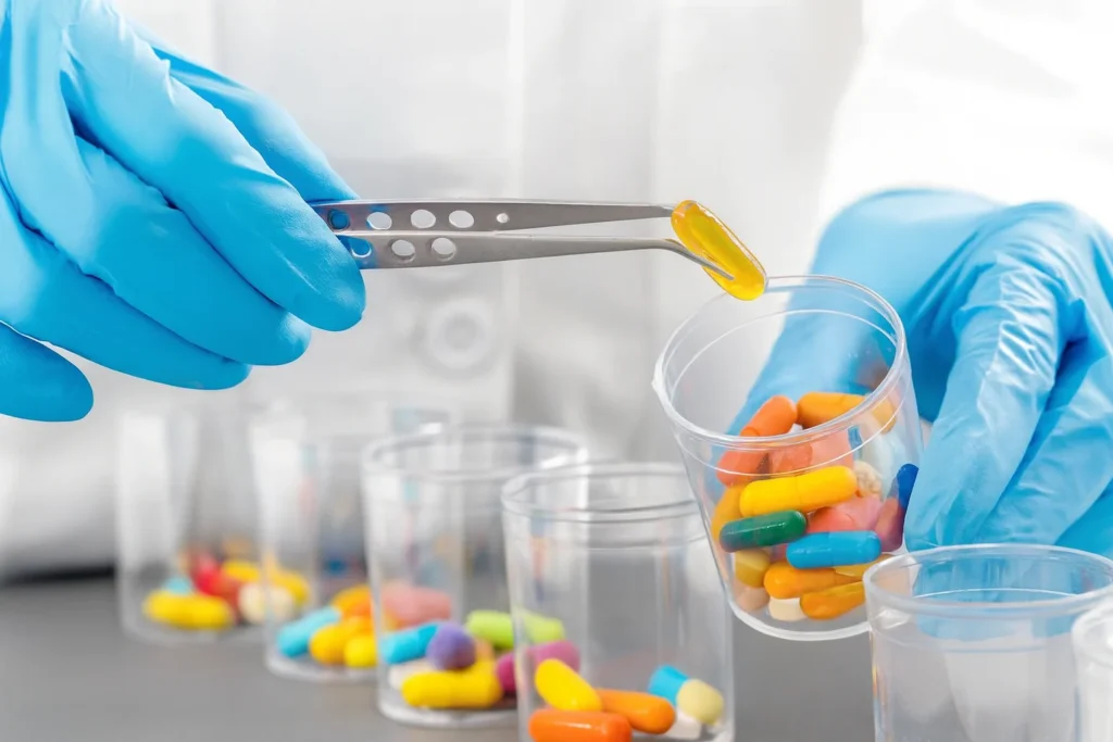 A compounding pharmacy technician in blue gloves sorting various colorful medications with tweezers into clear cups.